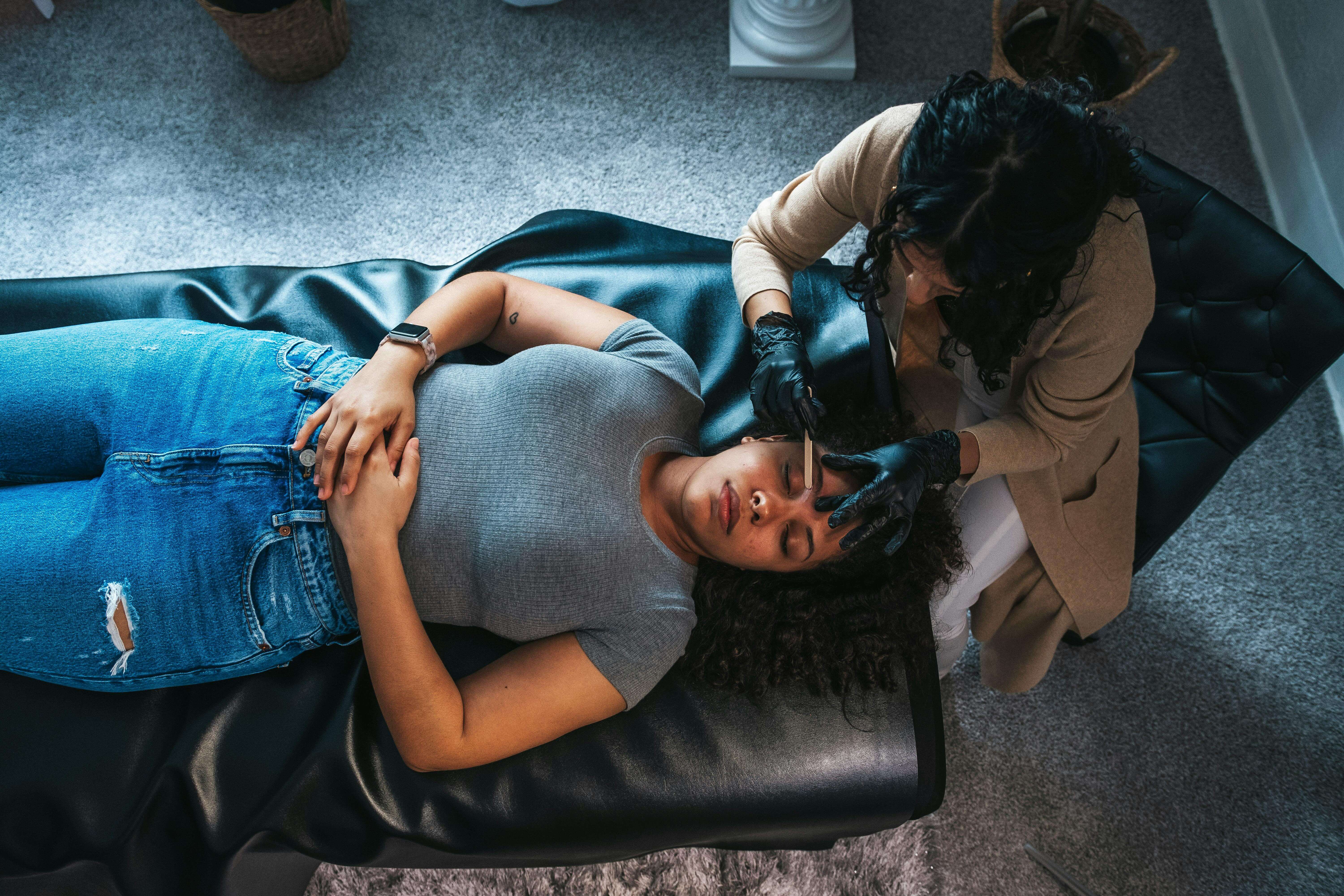 A woman receiving an eyebrow shaping service by an esthetician.